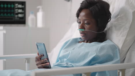 A-young-woman-writes-a-message-on-her-phone-while-lying-in-a-hospital-ward.-An-African-girl-is-lying-in-a-ward-connected-to-ECG-and-oxygen-devices-in-a-mask-and-writes-messages-to-relatives